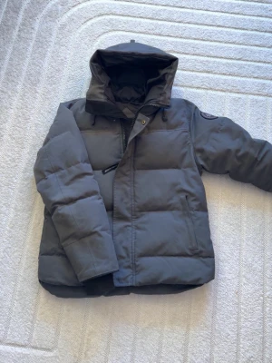 Mörkgrå dunjacka med huva - Säljer en mörkgrå dunjacka med stor, justerbar huva och dragkedja framtill. Jackan har två sidofickor och en patch på ärmen. Den är vadderad och perfekt för kalla vinterdagar. Passar dig som vill ha en stilren och varm jacka. Funkar även för storlek M