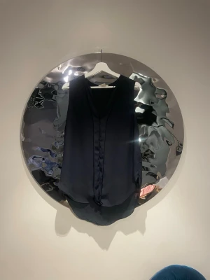 Svart ärmlös blus  - Stilren svart blus i ett lätt, flowy material med v-ringning och dekorativ knytning framtill. Ärmlös modell med något längre bakstycke för en avslappnad look. Perfekt att matcha med jeans eller kjol för en trendig outfit.