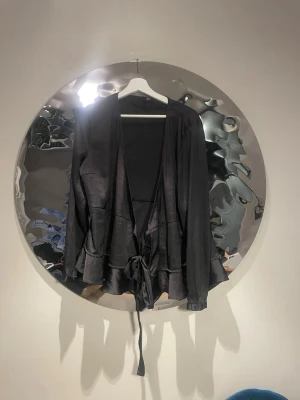 Svart omlottkofta med volang - Snygg svart omlottkofta med volangkant och sleek finish. Koftan har  independent ärmar och knyts i midjan med ett band för en schysst siluett. Perfekt för lager-på-lager och funkar till det mesta. Materialet känns lätt och har en lite glansig yta.