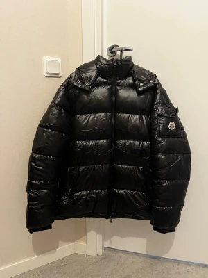 Svart pufferjacka från Moncler - Säljer en svart pufferjacka från Moncler med hög krage och dragkedja framtill. Jackan har Moncler-logga på ärmen, quiltad design och flera fickor. Insidan har ett färgglatt serietryck. Perfekt för kalla vinterdagar och riktigt snygg streetstil.
