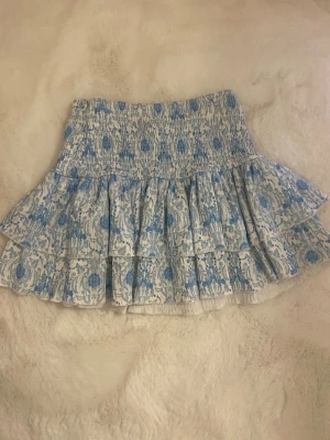 Blåvit volangkjol från RYVLS - Supersöt kortkjol från RYVLS i blått och vitt med volanger och smock i midjan. Kjolen har ett blommigt mönster och är gjord i ett lätt och luftigt bomullstyg, perfekt för varma dagar. Passar dig som gillar en feminin och trendig stil.