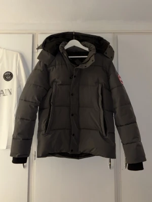 Mörkgrå dunjacka från Canada Goose - Snygg mörkgrå dunjacka från Canada Goose med klassisk patch på ärmen. Jackan har huva, dragkedja och knappar framtill samt två stora fickor. Perfekt för kalla vinterdagar och riktigt varm tack vare dunfyllningen.