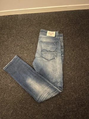 Blå slim fit jeans från Replay - Säljer ett par blå slim fit jeans från Replay med snyggt slitna detaljer och klassisk femficksmodell. Jeansen har en ljus tvätt framtill och bak, samt Replay-lapp bak i midjan. Perfekta för en avslappnad och trendig look.