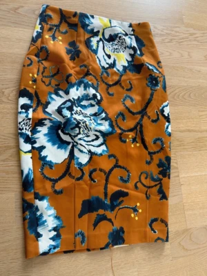 Blommig pennkjol i orange och blått - Snygg pennkjol med stora blommor i blått, vitt och gult på en orange bakgrund. Kjolen har en figurnära passform och är perfekt för dig som vill sticka ut med färg och mönster. Materialet känns slätt och bekvämt.