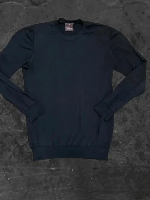 Svart tröja från Oscar Jacobson - Klassisk svart tröja från Oscar Jacobson i storlek M. Tröjan har rund halsringning, långa ärmar och ribbade muddar vid ärmslut och nederkant. Perfekt för en stilren och enkel look.