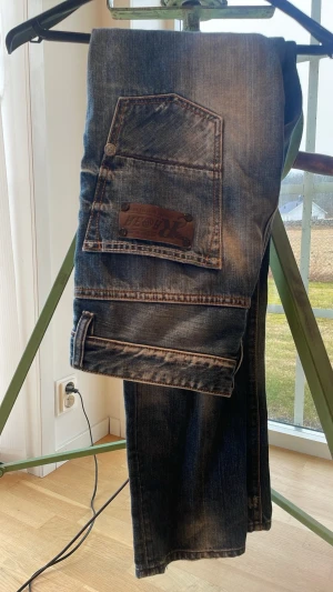 Blå raka jeans från Ecko Unltd - Säljer ett par blå jeans från Ecko Unltd med raka ben och slitna detaljer. Jeansen har klassisk femficksdesign, tydliga slitningar och en stor bakficka med läderpatch. Perfekta för en avslappnad streetstil. Strl 31/32