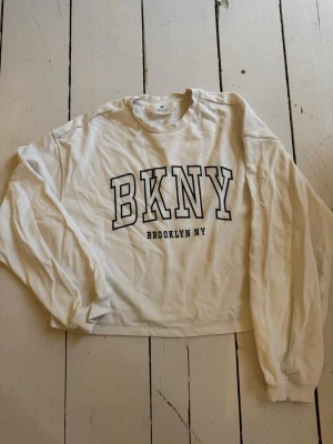 Vit sweatshirt med BKNY-tryck - Vit sweatshirt med svart BKNY-tryck och texten Brooklyn NY på bröstet. Tröjan har rund hals, långa ärmar och ribbade muddar. Perfekt för en avslappnad streetwear-look.