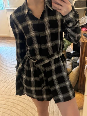 Svartvit rutig skjortklänning med bälte - Svart och vit rutig skjortklänning med lång ärm och knytbälte i midjan. Klänningen har klassisk krage och är kort i modellen. Perfekt för en trendig och avslappnad look.