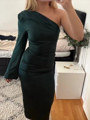 Mörkgrön oneshoulder långklänning - Snygg mörkgrön långklänning med oneshoulder-design och lång ärm på ena sidan. Klänningen har en slits baktill, en dragkedja i sidan och på axeln finns en axelvadd. Från gina tricot i strl 34. Bara använd den enda gång alltså så gott som ny