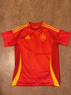 Spaniens matchtröja - Säljer en röd matchtröja från Adidas med Spaniens landslagsmärke på bröstet. Tröjan har gula detaljer och klassiska Adidas-ränder på axlarna. Tillverkad i lätt och ventilerande material, perfekt för fotboll eller träning.