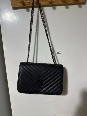 Svart axelväska från Saint Laurent - Säljer en svart axelväska från Saint Laurent med kedjerem och quiltat mönster i chevron-stil. Väskan har YSL-logga framtill och är tillverkad i slätt läder. Perfekt för dig som gillar stilrena och lyxiga accessoarer.