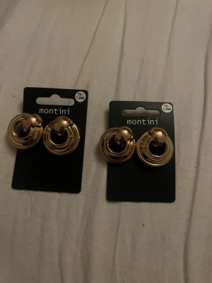 Guldiga statementörhängen från Montini - Säljer två par guldiga statementörhängen från Montini. Örhängena har en rund, tredimensionell design med cirkelformade ringar och en klotformad detalj upptill. Perfekta för dig som gillar trendiga accessoarer i metall-look. Jag säljer 1 för 80kr och 2 för 180kr