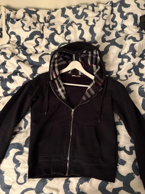 Svart ziphoodie från Burberry - Väldigt mysig och fräsch svart hoodie från Burberry med klassiskt rutigt foder i huvan och dragkedja framtill. Justerbar huva, 9/10 skick, använt ca 4-5 gånger. Storlek M sitter som en S/M. Hör av er vid intresse, priset sänkt med 250 kr redan!!!