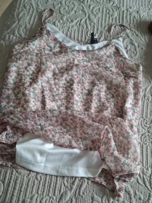 Blommig linnetopp från Vero Moda - Söt linnetopp från Vero moda med tunna axelband och dubbla lager. Toppen har ett blommigt mönster i ljusrosa, grön och vit på ett skirt yttertyg, samt ett vitt innerfoder. Perfekt för sommarens varma dagar.