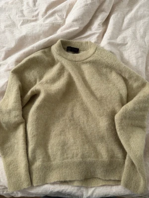 Gul/grön stickad tröja i ullmix - Mysig beige stickad tröja med rund hals och ribbade muddar. Tröjan har en avslappnad passform och är tillverkad i en mjuk ullblandning. 