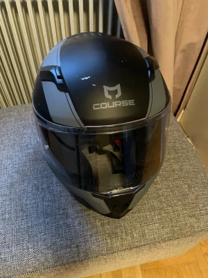Svart och grå motorcykel mentalm från Course - Snygg svart och grå hjälm från Course med visir som kan fällas upp. Hjälmen har en modern design med matta och blanka detaljer samt Course-loggan inpräntad framtill. Tillverkad i tålig plast och har vadderad insnog för extra komfort.