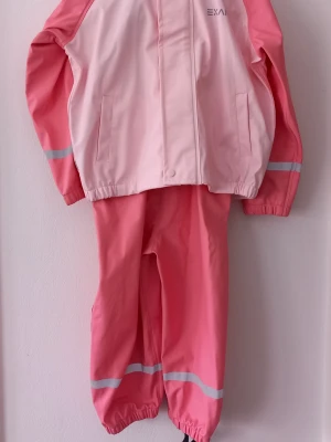 Barn - Rosa regnjacka från Exani - Storlek 98 - Säljer en snygg barn rosa regnjacka från Exani med ljusrosa detaljer och reflexband på ärmarna. Jackan är vattentät och har två fickor framtill. Perfekt för regniga dagar och håller dig torr. Materialet är slitstarkt och lätt att rengöra. Storlek 98 i barnkläder motsvarar generellt en ålder på 2–3 år. Storleken är baserad på barnets längd i centimeter (centilong), vilket innebär att den passar bäst för barn som är cirka 98 cm långa. Det är en vanlig storlek för barn på förskola. 