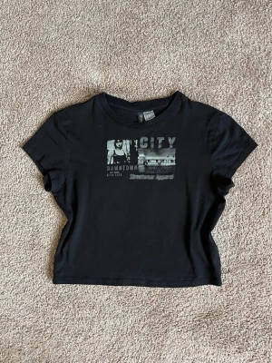 Svart croppad t-shirt med citytryck - Snygg svart croppad t-shirt med coolt citytryck framtill. T-shirten har korta ärmar och rund halsringning. Perfekt för dig som gillar streetstyle och vill ha en enkel men trendig look.