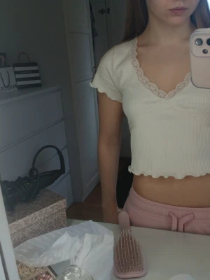 Vit croppad topp med spetskant - Söt vit croppad topp med korta ärmar och v-ringning. Toppen har en dekorativ spetskant runt halsen och vågig kant nertill. Perfekt för en avslappnad och trendig look.