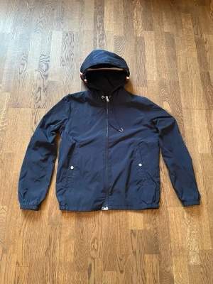 Mörkblå jacka - Snygg mörkblå jacka från Moncler med huva och dragkedja framtill. Jackan har två framfickor med knappar och en justerbar dragsko i huvan. På huvan finns en randig detalj i vitt och rött. Stl 1 motsvarar storlek S. 