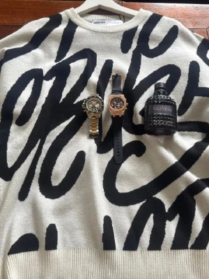 Vit och svart stickad tröja Bershka - Snygg stickad tröja från Bershka i vit bas med stora svarta grafiska bokstäver. Rund halsringning och ribbade muddar. Perfekt för dig som gillar trendiga och unika plagg med streetwear-vibe. Materialet är mjukt och bekvämt.