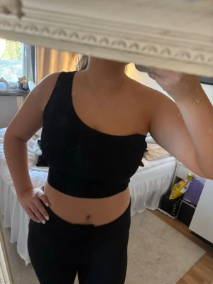 Svart ribbad oneshoulder topp - Säljer en svart ribbad oneshoulder topp med croppad passform. Toppen är ärmlös och har en snygg asymmetrisk design som ger en trendig look. Perfekt att matcha med höga jeans eller kjol.