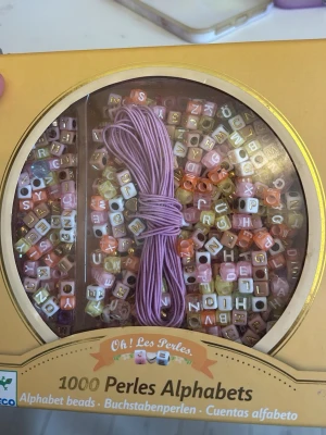 Pärlset med alfabetspärlor och snöre - Säljer ett pärlset från Oh! Les Perles med 1000 alfabetspärlor i olika färger som rosa, guld, vitt och orange. Setet innehåller även ett lila snöre för att skapa egna armband med personliga texter. Perfekt för dig som vill designa egna smycken med bokstavspärlor.