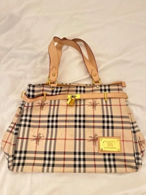 Burberry handväska - Snygg handväska från Burberry med klassiskt beige, svart och rött rutmönster. Väskan har ljusbruna handtag, guldiga metalldetaljer och en guldfärgad logoplatta framtill🫶🏼