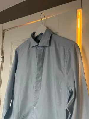 Ljusblå skjorta från Riley Slim Fit - Säljer en stilren ljusblå skjorta från Riley i slim fit-modell. Skjortan har klassisk krage, knappar framtill och är långärmad. Perfekt för dig som gillar en clean och enkel look.