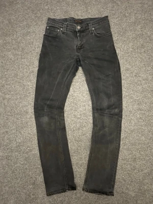 Svarta slim fit jeans från Nudie Jeans - Säljer ett par svarta slim fit jeans från Nudie Jeans med klassisk femficksmodell och diskreta slitningar. Jeansen har en smal passform, normal midja och är tillverkade i stretchig denim för extra komfort. Snygga detaljer på bakfickorna.