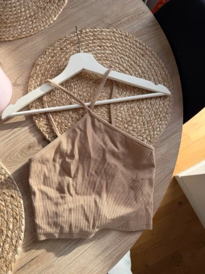 Beige ribbad croppad linnetopp - Säljer en beige croppad linnetopp med smala axelband som korsas i ryggen. Toppen är ribbad och har en enkel, stilren design som passar perfekt till sommaren. Materialet känns mjukt och stretchigt för en bekväm passform.