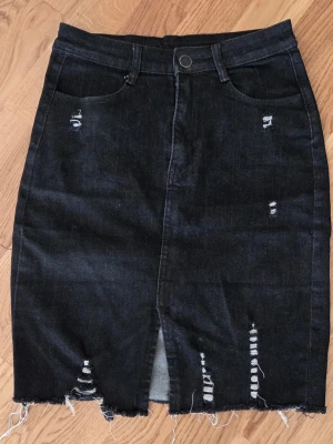 Svart jeanskjol med slitningar Stl S - Snygg svart jeanskjol med slitna detaljer och rå kant nertill. STL S Kjolen har fem fickor, bälteshällor och en liten slits framtill. Perfekt för en trendig och avslappnad stil Längd 48 cm Midjemått 31 cm ( 62 cm ) runt 75 % bomull 23 % polyester 2 % elastan Anv 1 - 2 ggr