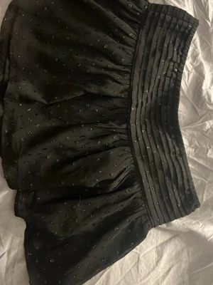 Svart kortkjol med pärlor från Gina Tricot - Supersnygg svart kortkjol från Gina Tricot med glittriga pärldetaljer och bred, veckad midja. Kjolen har en lätt glansig yta och är perfekt för dig som vill sticka ut lite extra. Passar till många olika stilar och är enkel att matcha.