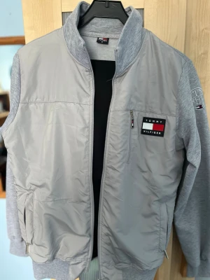 Grå ziptröja från Tommy Hilfiger  - Snygg grå ziptröja från Tommy Hilfiger med broderad logga på bröstet och ärmen. Tröjan har blandat material med quiltad framsida och mjuka ärmar och rygg. Praktiska fickor med dragkedja och hög krage för extra stilpoäng. 