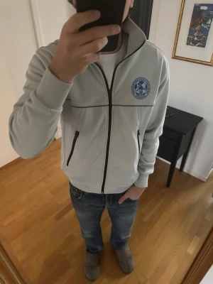 Ifk Göteborg Änglarna zip tröja - Vit ziptröja med svart dragkedja och detaljer, samt klubbmärke på bröstet och stor blå text 'Supporterklubben Änglarna' på ryggen. Tröjan har hög krage, ribbade muddar och två fickor med dragkedja. Perfekt för dig som vill visa ditt supporterskap.