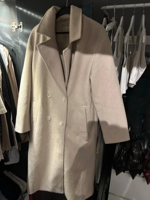 Beige ullkappa från Amisu - Stilren beige roadside ullkappa från Amisu Classic Collection. Kappan har dubbelknäppning framtill, två fick scarf och were bred krage. Perfekt ket höst och vinter för dig som vill ha en klassisk och tidlös look.