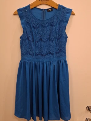 Blå spetsklänning från Oasis - Snygg blå kortklänning från Oasis med spetsdetaljer på överdelen och rund halsringning. Klänningen är ärmlös och har en markerad midja med ett lätt utställt kjolparti. Dragkedja bak för enkel påklädning. Perfekt för dig som vill ha en feminin look.