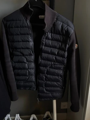 Svart dunjacka från Moncler - Snygg svart dunjacka från Moncler med quiltad front och ärmar i stickad kvalitet. Jackan har hög krage, dragkedja framtill och diskret Moncler-logga på ärmen. Perfekt för kalla dagar och riktigt stilren look.