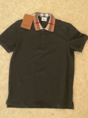 Svart Burberry polo - Snygg svart t-shirt från Burberry med klassisk rutig krage i beige, vitt, brunt och rött. T-shirten har korta ärmar, normal passform och tre knappar framtill. Tillverkad i mjuk bomull för en skön känsla.