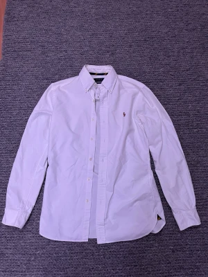 Vit skjorta från Polo Ralph Lauren - Klassisk vit skjorta från Polo Ralph Lauren med broderad logga på bröstet. Skjortan har lång ärm, knappar framtill och button-down krage. Perfekt för en clean och stilren look.