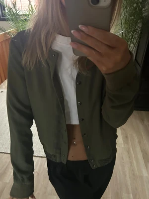 Olivgrön cardiganjacka från Vero Moda  - Snygg olivgrön cardiganjacka från Vero Moda med rund halsringning och tryckknappar framtill. Jackan har ribbade muddar vid ärmslut och nederkant, och är tillverkad i ett mjukt tyg som känns bekvämt mot huden.