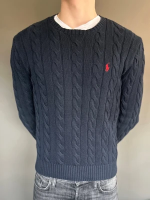 Mörkblå kabelstickad tröja Polo Ralph Lauren - Mörkblå kabelstickad tröja från Polo Ralph Lauren med rund halsringning och röd broderad logga på bröstet. Tröjan har ribbade muddar vid ärmslut och nederkant. Perfekt för en stilren och klassisk look. Nypris 2300kr+
