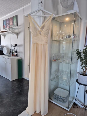 Lång ljus klänning med omlottband - långklänning i ljusgult med smala axelband. Klänningen har ett flytande fall och v-ringad urringning fram. Öppen rygg med bara korsade smala band.. Perfekt för speciella tillfällen när du vill känna dig extra fin.  Det lilla metallspännet vid dragkedjan sitter lite dåligt men påverkar inte passformen. 