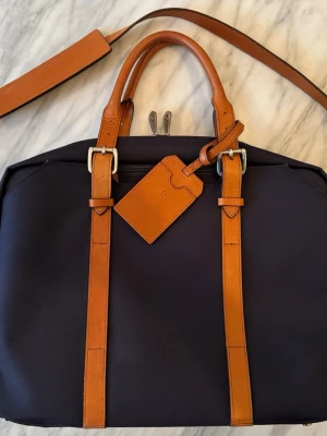 Portfölj från Stuart & Lau med läderdetaljer - Marinblå portfölj med handtag och axelrem i italienskt läder från Stuart & Lau. Väskan är i twill med vattentät baksida. Avtagbart datorfodral, praktiska fack och möjlighet att fästa på resväska. Använd ett fåtal gånger och i så gott som nyskick. Nypris 3500kr. 