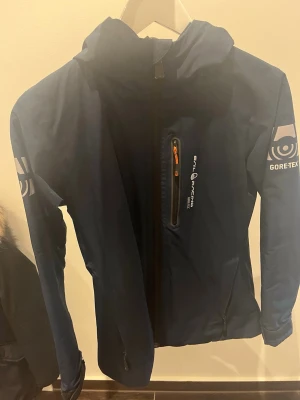 Blå Sail Racing Gore-Tex vindjacka - Snygg blå vindjacka från Sail Racing med Gore-Tex-material. Jackan har huva, dragkedja framtill, en bröstficka med orange detalj och loggor på ärm och rygg. Perfekt för blåsiga dagar och sportiga aktiviteter.