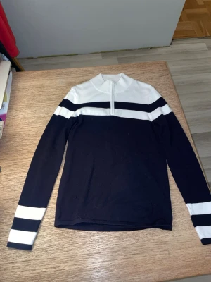 Svartvit stickad tröja med dragkedja från Jean Paul - Stickad tröja i svart och vitt med halv dragkedja framtill. Tröjan har långa ärmar och breda vita och svarta ränder på ärmar och över bröstet. Hög krage och rak passform, perfekt för en stilren look.