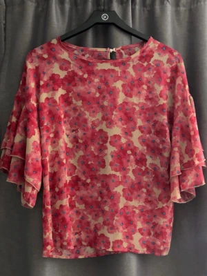 Blommig blus med volangärmar - Blus med färgglatt blommönster i rött, rosa och beige samt små blå detaljer. Modellen har rund halsringning och vida, volangiga trekvartsärmar som ger en luftig känsla. Materialet ser ut att vara lätt och något transparent, perfekt för en trendig look.