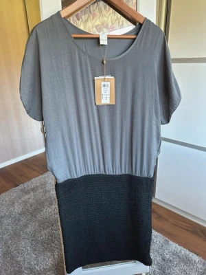 Grå och svart klänning från Vila - Tvåfärgad kl soppskuren klänning från Vila i storlek M. Överdelen är i mjukt grått tyg med rund halsringning och col korta ärmar, medan nederdelen är svart och har en us da stickad struktur. Enkel och stilren design som passar till många tillfällen.