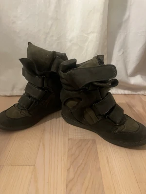 Mörkgröna höga sneakers med kardborre - Uppskattar storleken som 36 mer eller mindre. Det syns tyvärr inte vad det är för Stl se nedan, Köpte dessa och ett par beiga (se i profil) på loppis, storleken står ej på skorna därav har jag inte kunnat använda dom då de är på tok för små, lämna gärna prisförslag.  Skor med hög sula.Unika mörkgröna sneakers med högt skaft och flera breda kardborreband. Tillverkade i mocka och textil med vadderad plös och krage för extra komfort. Snygg streetstil som sticker ut och passar perfekt till jeans ell