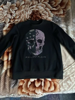 Philipp Plein - Snygg svart hoodie från Philipp Plein med ett stort döskallemotiv framtill, där ena halvan är täckt av glittrande stenar och den andra har en 3D-effekt med geometriska former. Tröjan har ribbade muddar och rund hals. Perfekt för dig som gillar edgy streetwear.